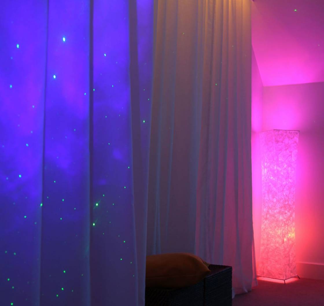 Image a room with curtains which are lit up blue and a lamp emitting pink and orange light in the corner