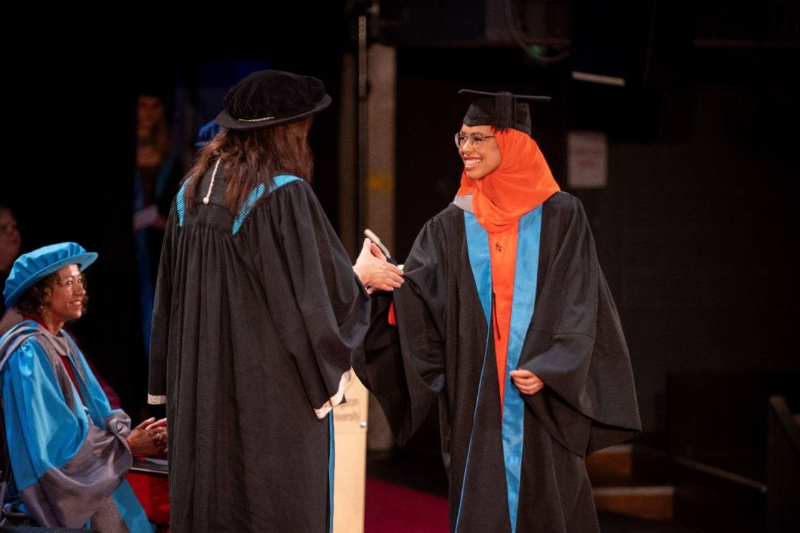 A smiling graduate shakes hand with professor while receiving her award on stage at a graduation ceremony