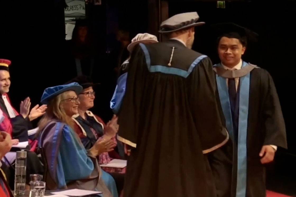 A smiling graduate shakes hand with professor while receiving her award on stage at a graduation ceremony