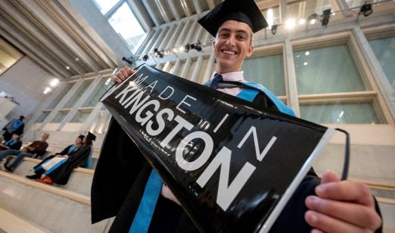 Smiling graduate in hat and gown, holding a banner that reads: Made in Kingston.