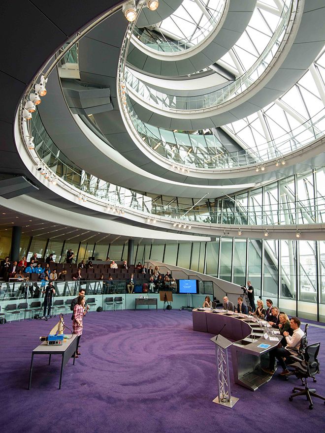 Two contestants pitch to a panel of judges with purple carpet and winding stairs and windows in the background