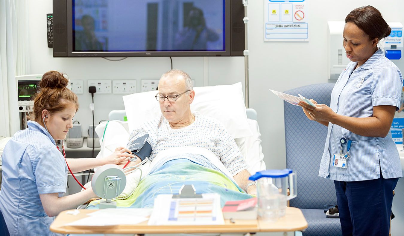 Picture of two nursing students in the University\'s skills labs with a pretend patient in the hospital bed.