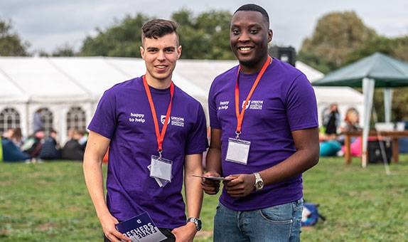 Two Union of Kingston Students' staff in purple T shirts with a marquee behind them