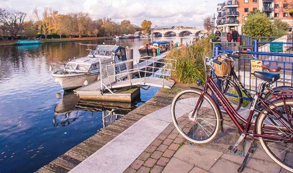 A bicycle parked on Kingston riverside showing Kingston bridge, boats and a wooded area on the other side of the river. 