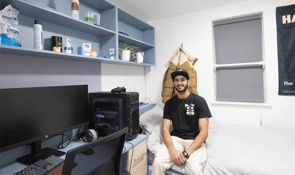 A male student sitting on their bed, next to the desk, in the smaller standard en suite room