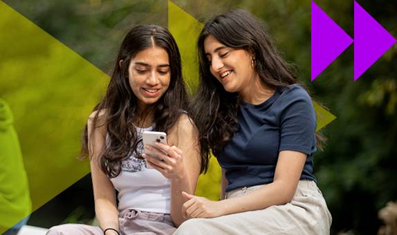 Two smiling female students looking at a phone, sitting on a wall with trees in the background