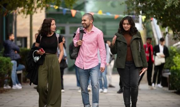 Three students walking together down a paved area of Kingston Hill campus. Bunting, trees and other students are in the background.