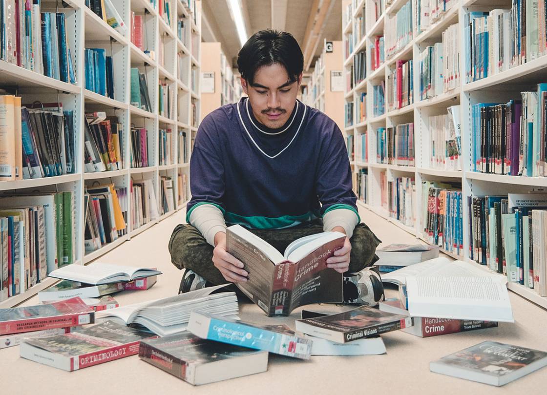 A male student sitting on the library floor, reading while surrounded by books