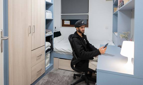 A male student sitting at his desk next to the bed in the standard en suite room in Chancellors Hall 