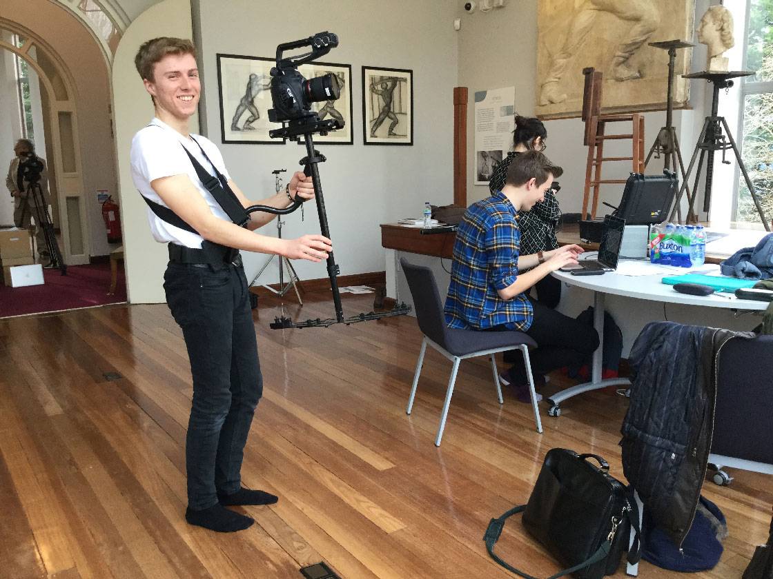 A student holding camera equipment, standing next to a table where two students work on laptops.