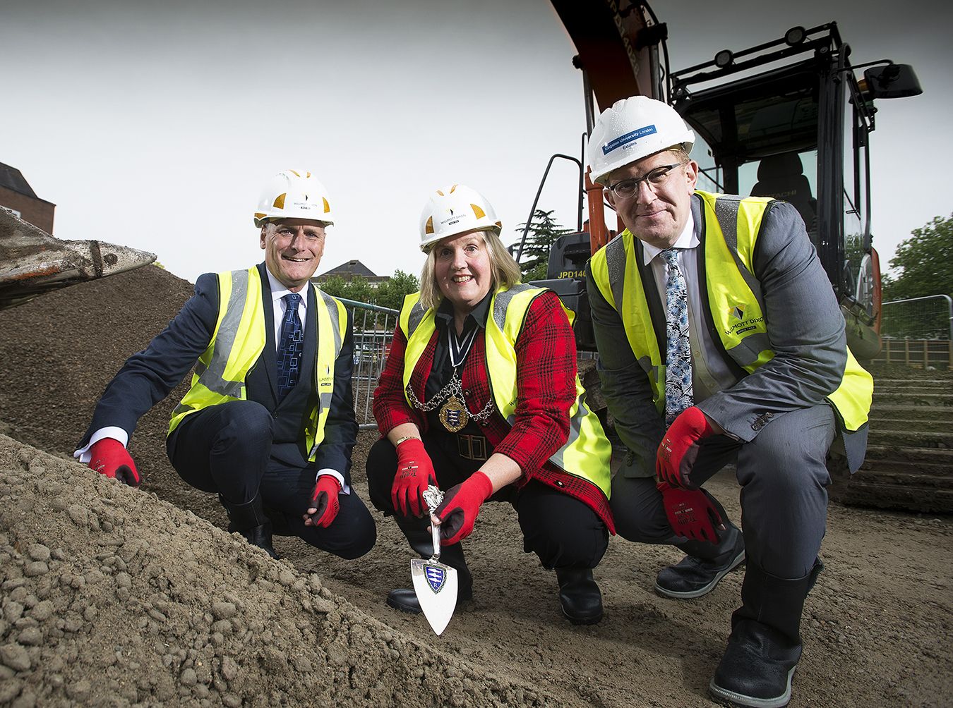 Photo os two men and one woman wearing high-vis jackets and hard hats with the woman holding a silver trowel.