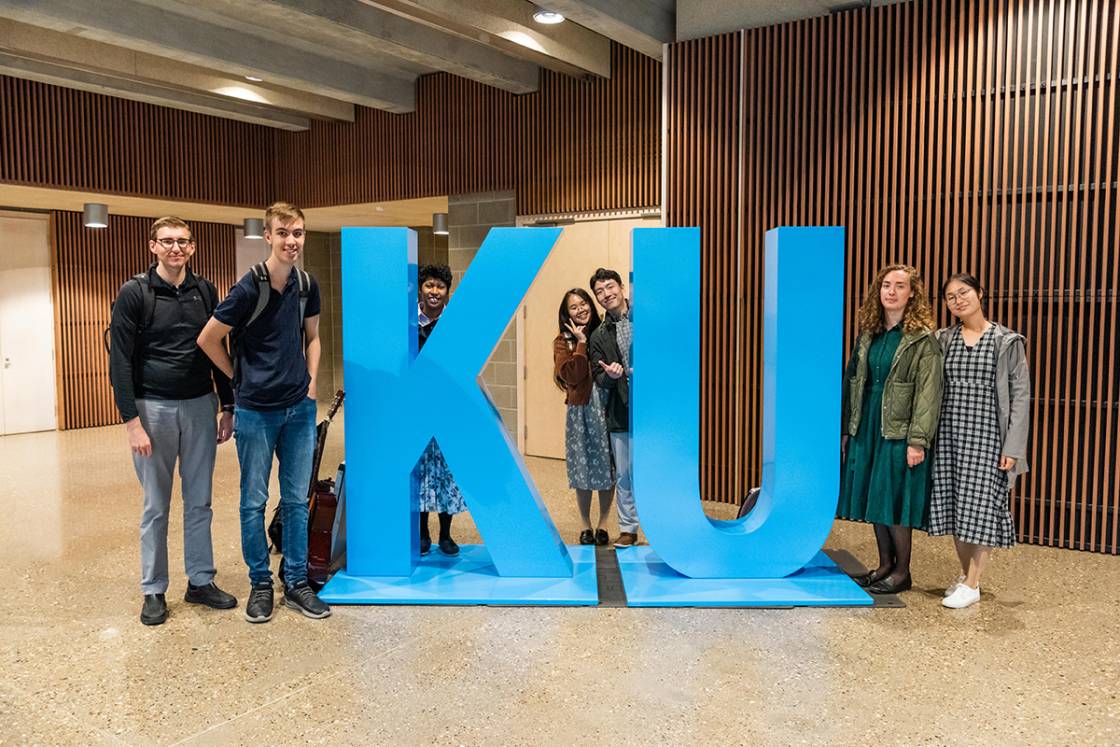 Seven happy students standing with a big blue statue that says "KU"