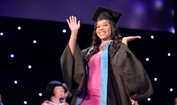 Smiling graduate on stage, waving and wearing hat and gown